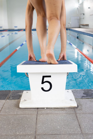 Biracial young female swimmer standing on starting block indoors, ready to dive. She has light brown skin, dark hair, wearing swimsuit, unaltered.の写真素材