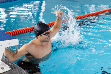 Caucasian young male swimmer celebrating indoors in pool, splashing water. He has short dark hair,...