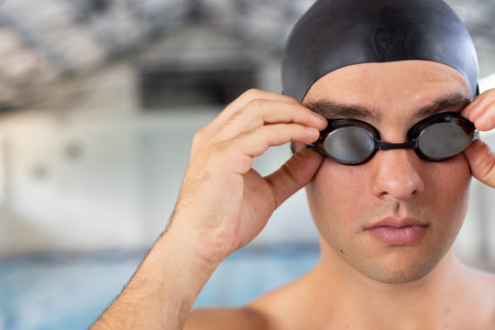 Caucasian young male swimmer adjusting goggles indoors, standing by pool, copy space. He has short brown hair, fair skin, and is wearing a black swim capの写真素材