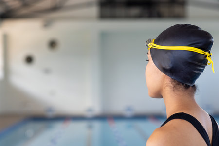 Biracial young female swimmer wearing a black cap is looking at a pool indoors, copy space. She has dark hair tied back, brown eyes, and is wearing a swimsuit, unalteredの写真素材