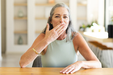 At home, mature Caucasian woman covering mouth during video call. Sporting silver hair and light...