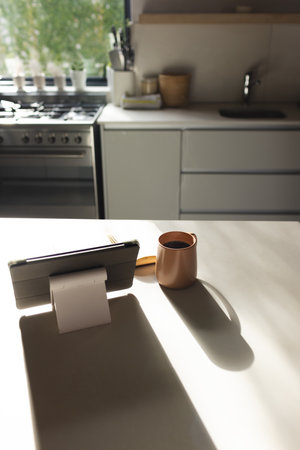 Sunlight shining on kitchen counter with cup and tablet at home with copy space. Warm light creates...
