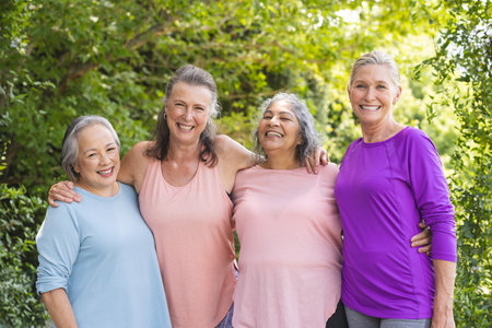 Outdoors, senior female friends standing together, sharing smiles. Dressed casually with blue...