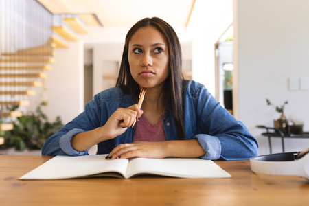 Biracial woman holds pencil, looks thoughtful on video call at home. Dressed casually, with long...