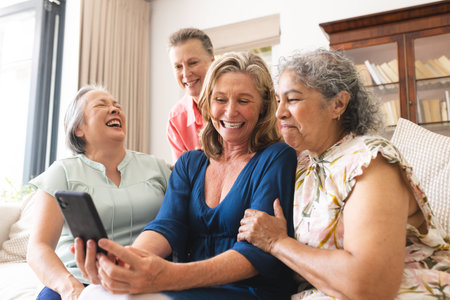 Diverse senior female friends laughing at home, looking at tablet. Wearing casual clothes, sharing...