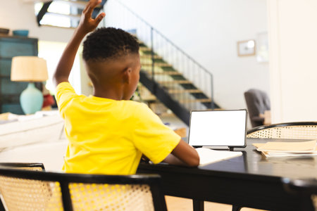 African American boy sitting at table, raising hand near tablet at home with copy space. He has short hair, wearing a yellow shirt, focused on a blank screen, unaltered.の写真素材