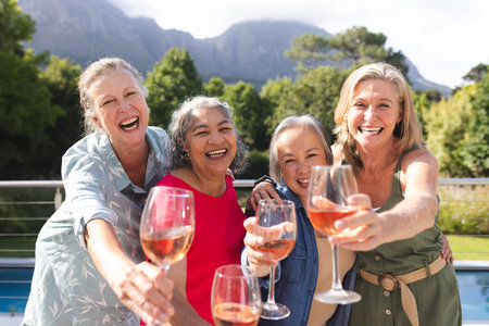 Diverse senior female friends holding wine glasses, laughing outdoors. Wearing casual clothes, enjoying sunny day with a blue background, unalteredの写真素材