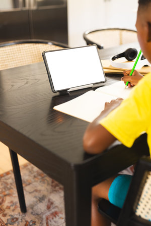 African American boy in yellow shirt writing in notebook using tablet, copy space. Wearing...