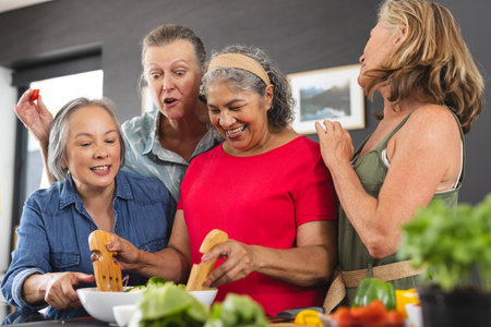 Diverse senior female friends cooking together at home. Wearing casual clothes, sharing joyful...