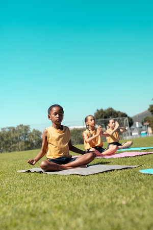 Composite of diverse schoolgirls meditating while sitting on mats over grassy field under clear sky. Copy space, yoga, zen, exercise, fitness, childhood, nature, summer, together, german, school.の写真素材