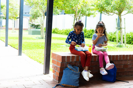 In a school courtyard, two young Hispanic girls enjoy a lunch break, copy space. They sit on a brick ledge, eating from bowls, with backpacks beside them, unalteredの写真素材