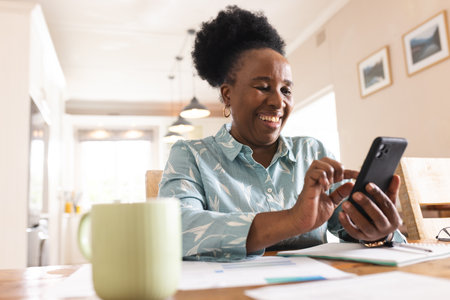 Happy senior african american woman with documents on table using smartphone at home. Domestic life, retirement, technology and lifestyle, unaltered.の写真素材