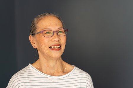 Smiling elderly asian woman with glasses looking up, wearing striped shirt. Senior, joyful,...
