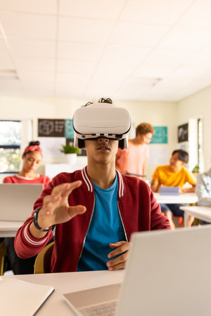 In high school, teenage boy using VR headset and laptop in classroom, exploring technology. Education, virtual reality, innovation, learning, immersive, digitalの写真素材