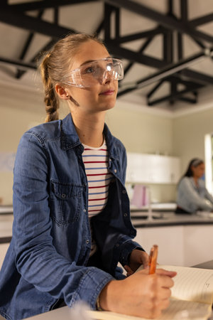 In high school, teenage girl wearing safety goggles writing notes in classroom. Education, learning, studying, teenager, science, laboratoryの写真素材