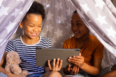 Under blanket fort, brother and sister playing together using tablet, smiling and enjoying time, in...