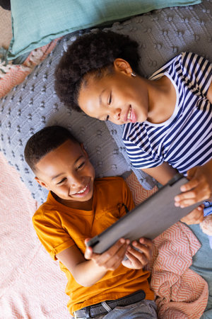 young brother and sister lying on bed, smiling and using tablet together at home. Kids, technology,...
