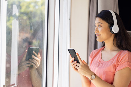 "Listening to music on headphones, Asian woman holding smartphone and looking outside, copy space. Technology, relaxation, leisure, streaming, lifestyle, enjoymentの写真素材