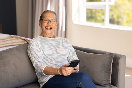 Smiling elderly Asian woman sitting on couch holding smartphone in living room. Senior, happiness,...
