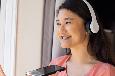 "Smiling Asian woman wearing headphones holding smartphone and looking out window. Technology, music, relaxation, indoors, lifestyle, listeningの写真素材