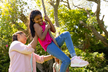 Mother helping daughter swing on rope in backyard, enjoying family time outdoors. bonding, playtime, happiness, childhood, parenting, recreationの写真素材