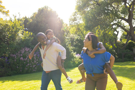 Playing in garden, multiracial parents giving piggyback rides to daughters, smiling and laughing....