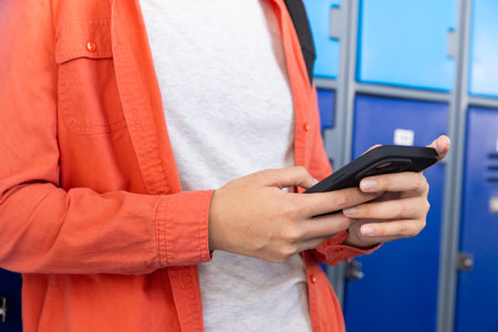 Using smartphone, teenage girl standing by lockers in school, wearing orange jacket. Education,...