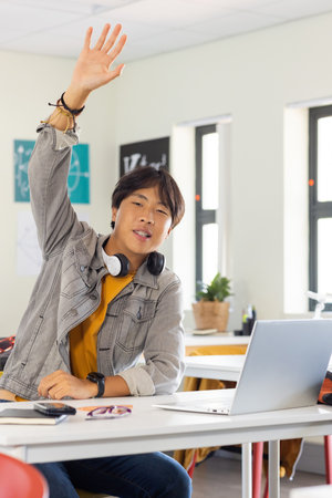 In school, asian teenage boy raising hand while sitting at desk with laptop. Education, learning, classroom, technology, studying, participationの写真素材