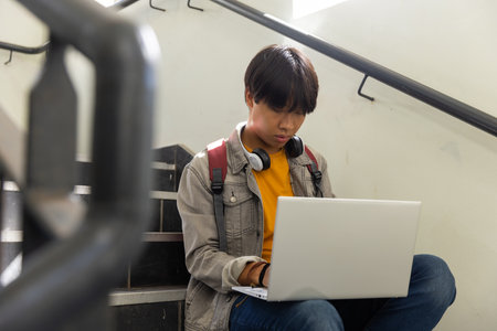 Asian teenage boy sitting on stairs using laptop and wearing headphones in school. education, technology, studying, learning, connectivityの写真素材