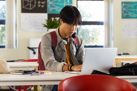 In school, asian teenage boy using laptop and studying in classroom with headphones. Education, learning, technology, academicの写真素材