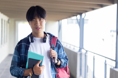 Carrying books and backpack, asian teenage boy standing in school hallway, looking confident. Education, confidence, learningの写真素材