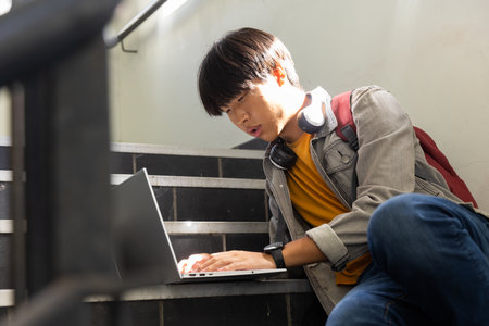 At school, Asian teenage boy sitting on stairs using laptop with headphones around neck. technology, studying, youth, education, asian teenage boy life, lifestyleの写真素材