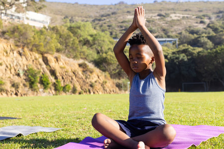 Practicing yoga, african american boy sitting on mat outdoors, meditating in peaceful setting. meditation, mindfulness, tranquility, wellness, relaxationの写真素材
