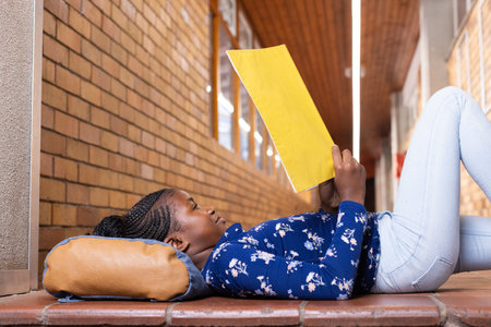 Reading notebook, african american girl lying on floor in school hallway, relaxing. Education, relaxation, student, learning, childhoodの写真素材
