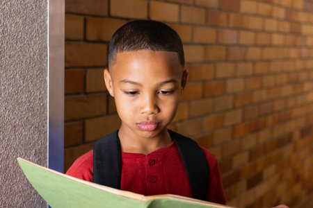 In school, boy reading notebook while standing against brick wall. Education, learning, studying, childhood, literature, studentの写真素材