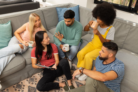 Group of young friends sitting on couch, drinking coffee, and talking together. Diverse friendship,...