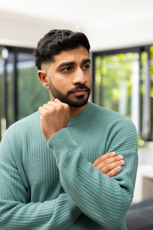 Thinking deeply, Indian man in green sweater standing with hand on chin. Contemplation, thoughtful, casual, pondering, concentration, decision-makingの写真素材