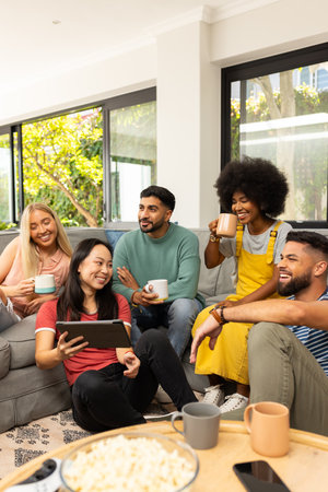 Diverse friends sitting on couch, drinking coffee, and watching tablet together, smiling....