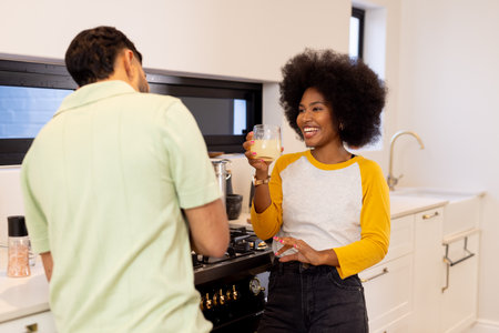 Smiling woman holding drink, talking with friend in modern kitchen. Friendship, happiness,...