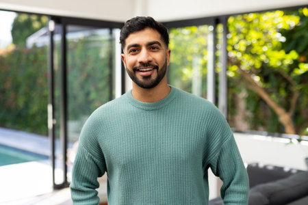 Smiling indian man in green sweater standing indoors near large windows. casual, male, bright,...