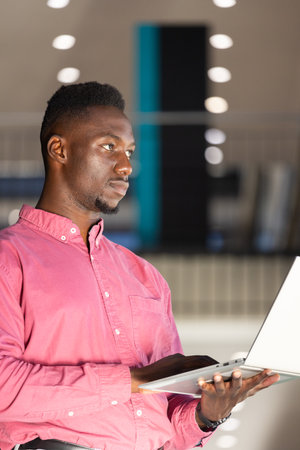 Using laptop, african american man in pink shirt focusing on work in modern office. business, technology, professional, concentration, indoor, workspaceの写真素材