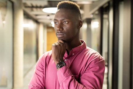 Thinking deeply, african american man in pink shirt standing in modern office hallway. Business,...