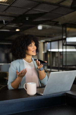 Speaking into smartphone, woman working on laptop with coffee mug in office. Business, technology, multitasking, productivity, workspace, remote workingの写真素材