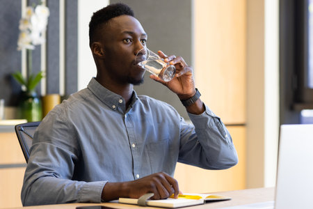 Drinking water, african american businessman sitting at desk with laptop and notepad in office. professionalism, hydration, workspace, productivity, notes, workingの写真素材