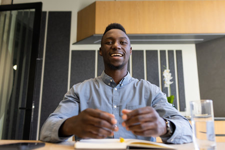 Smiling african american businessman sitting at desk with notebook and glass of water. office, professional, working, corporate, hydration, productivityの写真素材