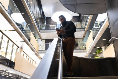 Businessman using smartphone while walking down stairs in modern office building, copy space. technology, professional, corporate, communication, urban, mobilityの写真素材