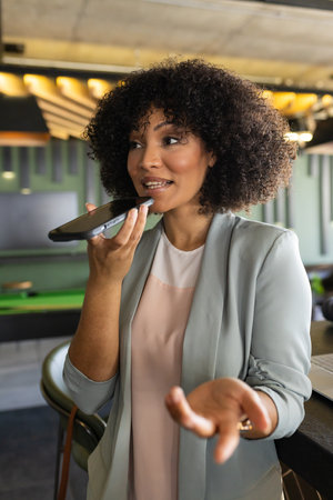 Speaking into smartphone, businesswoman using voice assistant in modern office. technology, communication, voice recognition, entrepreneur, productivity, professionalの写真素材