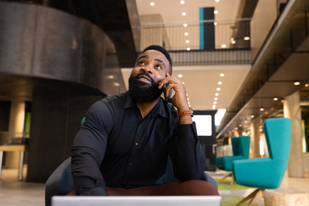 Talking on smartphone, African american man working on laptop in modern office setting. Business, technology, communication, workplace, professional, productivityの写真素材