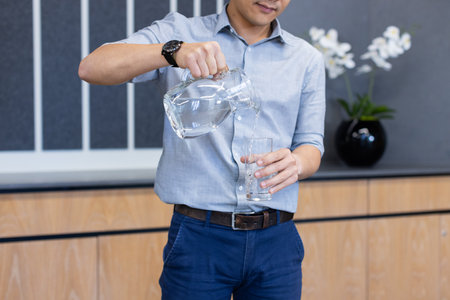 Pouring water from glass pitcher, Asian man in business attire hydrating. Hydration, refreshment, professional, drink, workplaceの写真素材