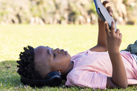 Lying on grass, african american boy using tablet and wearing headphones, enjoying outdoor learning. Technology, childhood, nature, relaxedの写真素材
